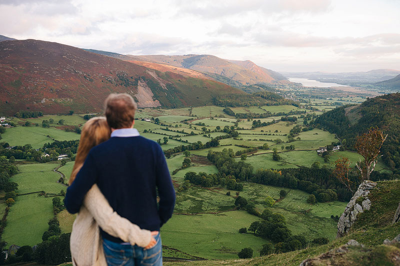 arms wrapped around each other, a couple look out from the top of the hill across the Lake District