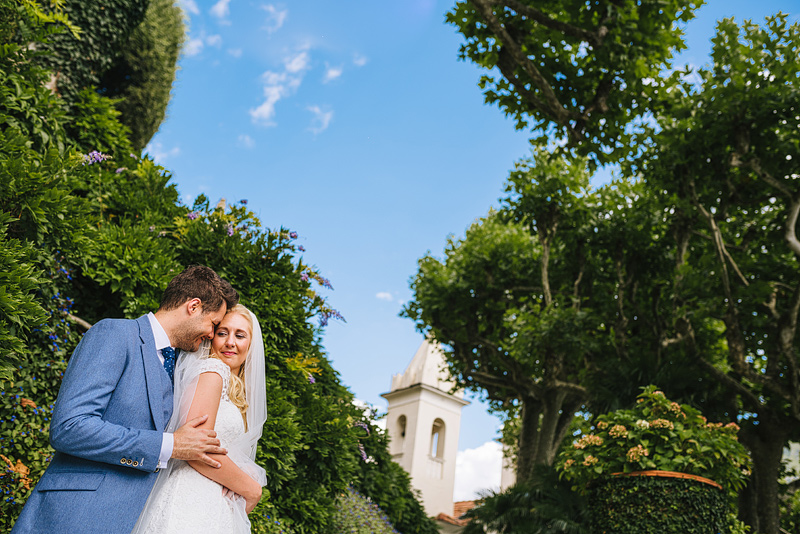 the couple embrace in the gardens with an old clock tower in the background
