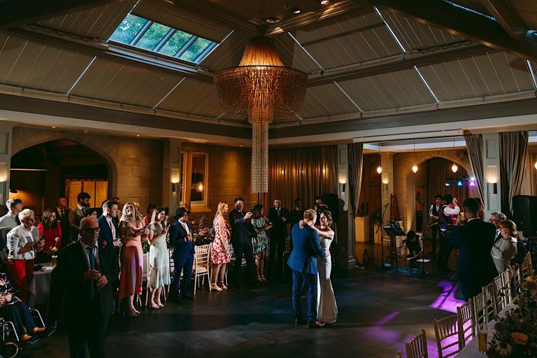 Lucy & Richard dancing at their wedding under the ornate golden chandelier in the courtyard room at Hampton Manor surrounded by their loved ones.